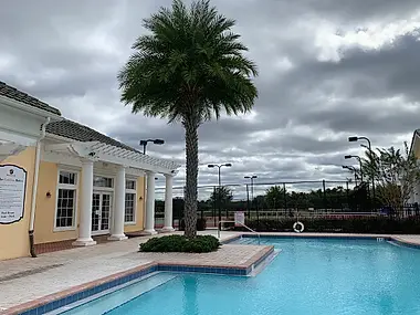 A clubhouse pool with tennis courts beyond, palm-shaded under a cloudy sky.
