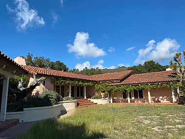 A single-story Spanish-style home with a terra cotta tile roof set against oak-covered hills.